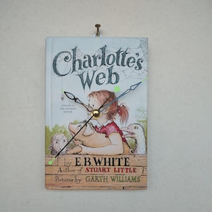 May include: A clock made from a book cover of "Charlotte's Web" by E.B. White. The clock face features the book's cover art with a girl, pig, and goose. The clock hands are black and silver, and the clock is mounted on a wall.
