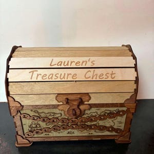 May include: A wooden treasure chest with a curved lid featuring the engraved text Lauren's Treasure Chest. The front panel displays a printed map design overlaid with a decorative wooden chain element and a central latch mechanism.