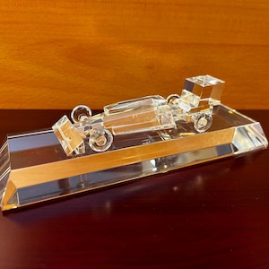 May include: A crystal figurine of a race car on a clear crystal base. The car is detailed with wheels and a spoiler.