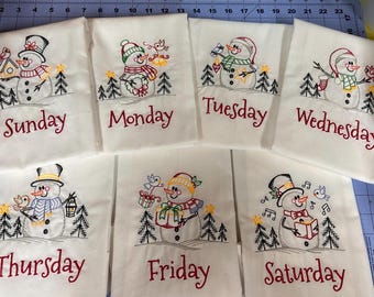 May include: Set of seven white tea towels with embroidered snowmen and the days of the week. Each towel features a different snowman design with a festive winter theme.