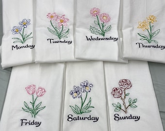 May include: Set of seven white tea towels with embroidered flowers and the days of the week. The flowers are embroidered in different colors, including pink, yellow, blue, and red. The days of the week are embroidered in black.