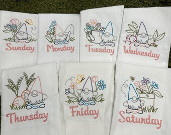 May include: Set of seven white embroidered kitchen towels, each featuring a gnome design with the days of the week in pink script. Gnomes are surrounded by flowers, butterflies, and garden elements. A charming and whimsical kitchen decor.