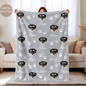 May include: A light gray fleece blanket featuring a repeating pattern of a black and tan dog's face and white paw prints. The name Rosie is printed in a white cursive script repeatedly across the fabric between the dog portraits.