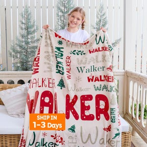 May include: A cream-colored blanket with the name "WALKER" in red, green, and gray lettering. Festive holiday-themed graphics, including a Christmas tree and snowflakes, are interspersed throughout the design. The blanket is held up by a person.