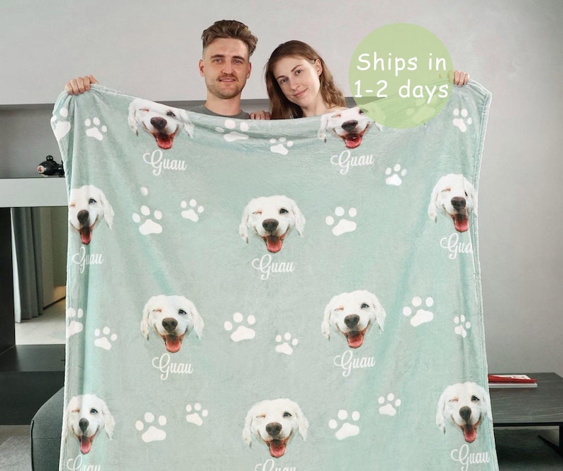 May include: A light green fleece blanket featuring a repeating pattern of white dog paw prints and the smiling face of a white dog with the text Guau printed in white script below each dog face across the entire surface of the fabric.