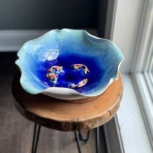 May include: A ceramic bowl with a blue and white glaze, featuring three small koi fish in the center. The bowl is sitting on a wooden stand.