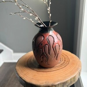 May include: A brown ceramic vase with a flame-like design, holding branches with small white buds. The vase sits on a round wooden side table with black metal legs. The vase is approximately 8 inches tall.