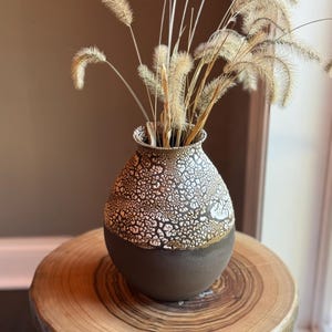 May include: A brown ceramic vase with a white crackle glaze, filled with dried beige grasses, sits on a wooden table.