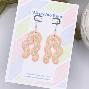 May include: A pair of peach-colored dangle earrings with a floral pattern. The earrings are made of polymer clay and have silver hooks. The earrings are displayed on a white card with the text "Waterloo Root" and social media information.