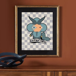 May include: Framed artwork featuring a cartoon Capricorn illustration with a blue and orange color scheme, set against a checkered background. The artwork is in a gold frame and includes the word "CAPRICORN".