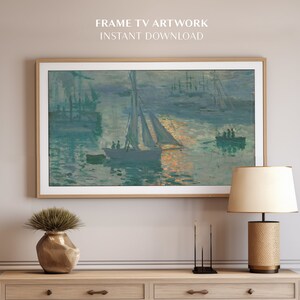 Op de afbeelding: Een ingelijst kunstwerk dat een zeezicht met zeilboten en een zonsondergang toont. Het kunstwerk wordt getoond aan een muur boven een houten dressoir met een lamp en een vaas met groen.