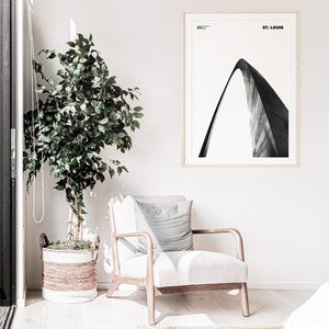 May include: A black and white photograph of the Gateway Arch in St. Louis, Missouri. The photograph is framed in a light wood frame and is hanging on a white wall. A potted plant and a chair are in the foreground.