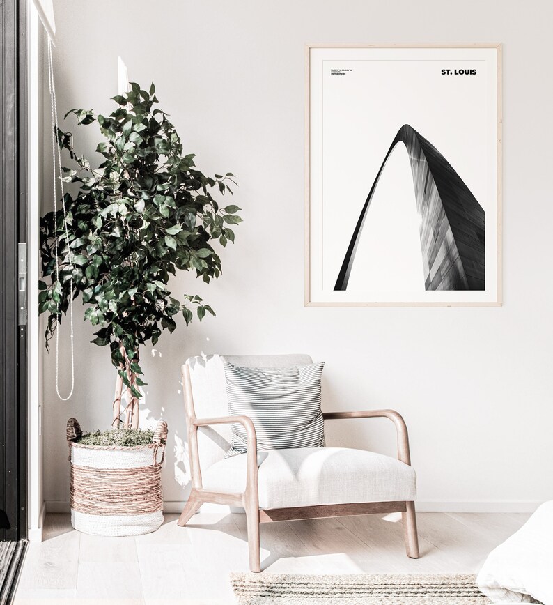 May include: A black and white photograph of the Gateway Arch in St. Louis, Missouri. The photograph is framed in a light wood frame and is hanging on a white wall. A potted plant and a chair are in the foreground.