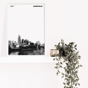 May include: Black and white photograph of the Nashville skyline with the text "NASHVILLE" in a white frame on a white wall. A potted plant hangs from a white shelf below the frame.