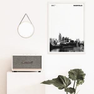 May include: A black and white photograph of the Nashville skyline with the text "NASHVILLE" in a white frame. A white speaker with a black grill and the word "Marshall" in gold lettering is on a white surface. A round mirror with a black leather strap is hanging on a white wall. A green plant is in the lower right corner of the image.