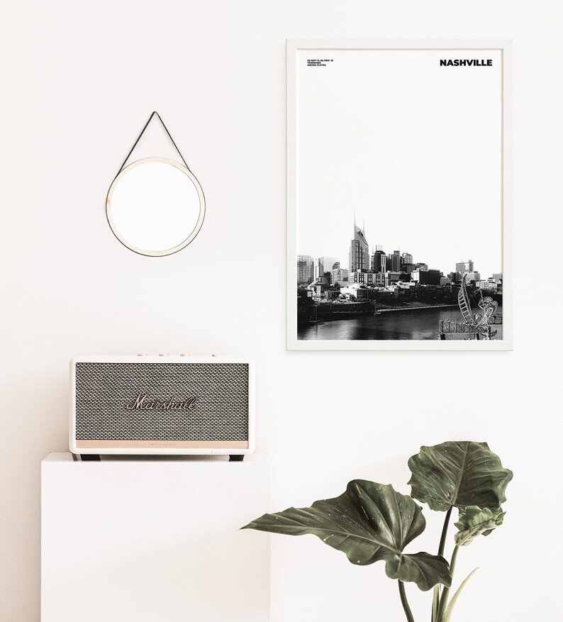 May include: A black and white photograph of the Nashville skyline with the text "NASHVILLE" in a white frame. A white speaker with a black grill and the word "Marshall" in gold lettering is on a white surface. A round mirror with a black leather strap is hanging on a white wall. A green plant is in the lower right corner of the image.