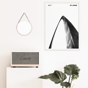May include: A black and white photograph of the Gateway Arch in St. Louis, Missouri. The photo is framed in a white frame and hangs on a white wall. A small round mirror with a black leather strap hangs above a white speaker.