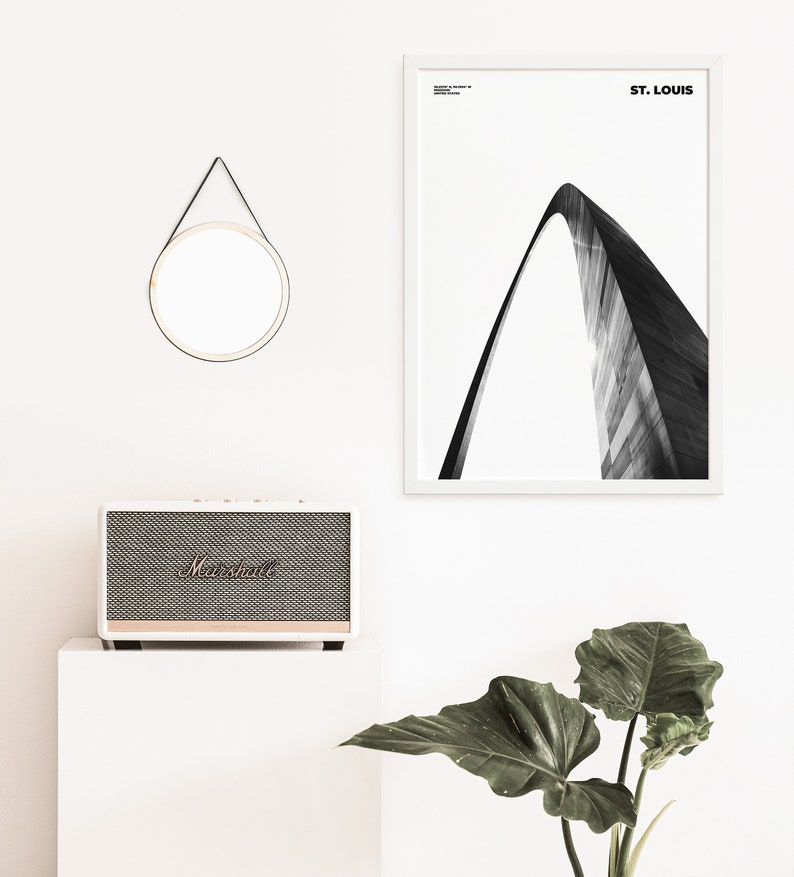 May include: A black and white photograph of the Gateway Arch in St. Louis, Missouri. The photo is framed in a white frame and hangs on a white wall. A small round mirror with a black leather strap hangs above a white speaker.