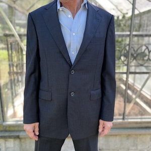 May include: A dark grey two-button suit with a light blue dress shirt. The jacket features a notched lapel and a chest pocket. The suit is paired with matching dark grey trousers. The man is standing in front of a greenhouse.