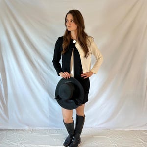 May include: A black and white jacket with a large white button. The jacket is worn over a white top and black skirt. The person is wearing black boots and holding a black hat.