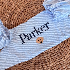 May include: Light blue and white striped baby romper with the name "Parker" embroidered in navy blue. A small tan dog face is embroidered below the name. The romper has puffed sleeves and snap closures.