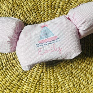 May include: Pink and white seersucker baby floatation device with a sailboat design embroidered on the front. The sailboat is light blue and pink, with the name "Emily" embroidered below. The floatation device is designed for babies.
