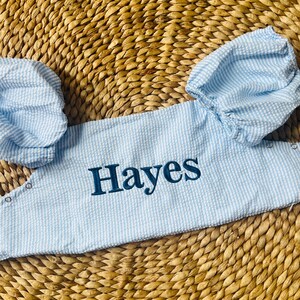 May include: Light blue seersucker baby romper with the name "Hayes" embroidered in navy blue. The romper features short sleeves and snaps for easy dressing. The fabric has a subtle striped pattern.