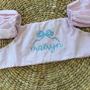 May include: Pink and white seersucker fabric top with puff sleeves. The top features a light blue embroidered bow and the name "maevyn" in cursive script. The garment has snap closures and is designed for a child.
