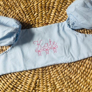 May include: Light blue seersucker bib with the name "Kate" embroidered in pink script, along with three pink bow accents. The bib features puffed sleeves and snap closures. The background is a woven, natural-toned surface.