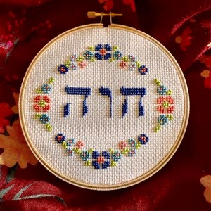 May include: A circular wooden embroidery hoop containing white Aida fabric with a cross-stitched design. The center features the Hebrew word YHWH in dark blue thread, surrounded by a circular wreath of small blue, red, and green flowers.