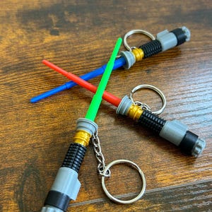 May include: Three lightsaber keychains with a silver, black, and gold design. The lightsabers are green, blue, and red. The keychains are on a wooden surface.