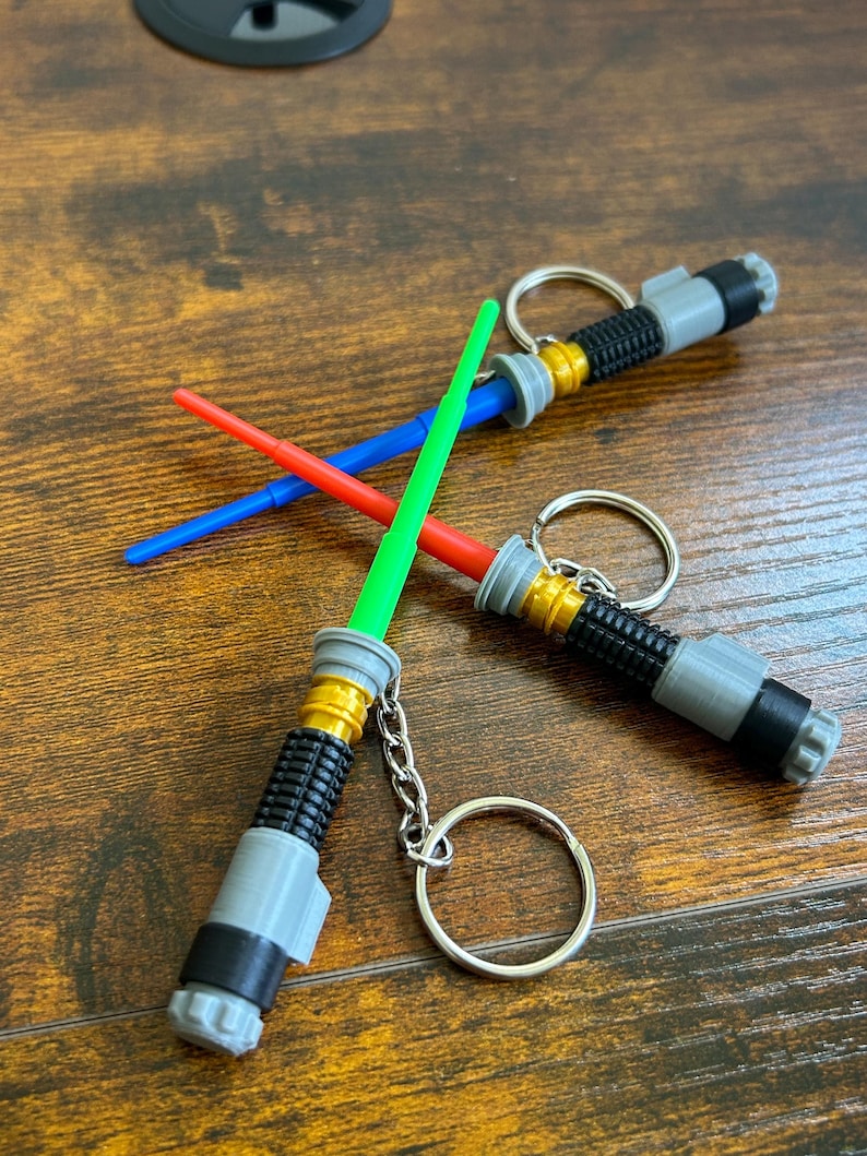May include: Three lightsaber keychains with a silver, black, and gold design. The lightsabers are green, blue, and red. The keychains are on a wooden surface.