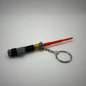 May include: A grey and gold lightsaber keyring with a red blade and a silver key ring.