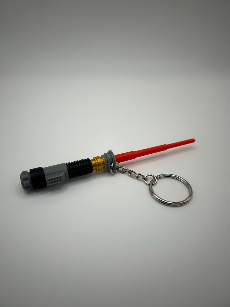 May include: A grey and gold lightsaber keyring with a red blade and a silver key ring.