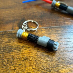May include: A grey and gold lightsaber keyring with a black band. The keyring is on a wooden surface.