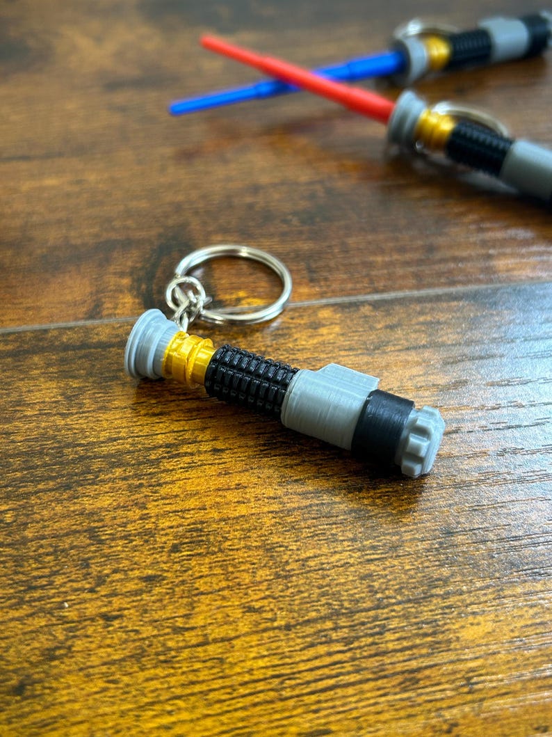 May include: A grey and gold lightsaber keyring with a black band. The keyring is on a wooden surface.