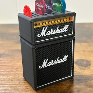 May include: A black and gold Marshall guitar amplifier with a speaker grill and a pick holder on top. The pick holder has five guitar picks in it, including a red pick with the text "Classic 70mm".
