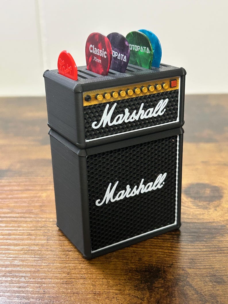 May include: A black and gold Marshall guitar amplifier with a speaker grill and a pick holder on top. The pick holder has five guitar picks in it, including a red pick with the text "Classic 70mm".