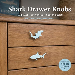 May include: Two light blue shark-shaped drawer knobs attached to a wooden dresser. The top knob is a hammerhead shark, and the bottom knob is a standard shark silhouette. Text on the image reads Shark Drawer Knobs, Handmade, 3D Printed, Custom Design, and Perfect for Ocean Lovers.