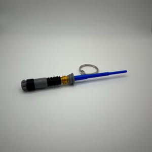May include: A blue lightsaber keyring with a grey and gold handle. The keyring has a silver ring.