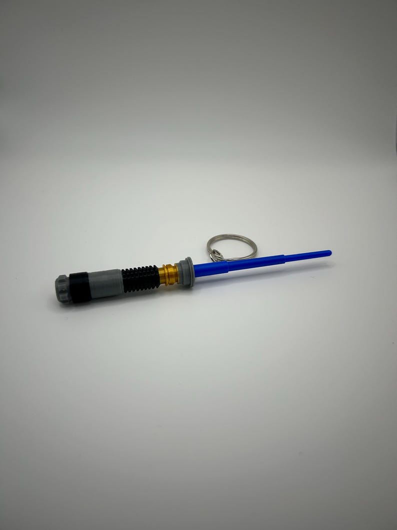 May include: A blue lightsaber keyring with a grey and gold handle. The keyring has a silver ring.