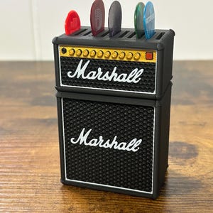 May include: Black and gold Marshall guitar pick holder shaped like a miniature amplifier. The holder has six slots for guitar picks and features the Marshall logo on the front.