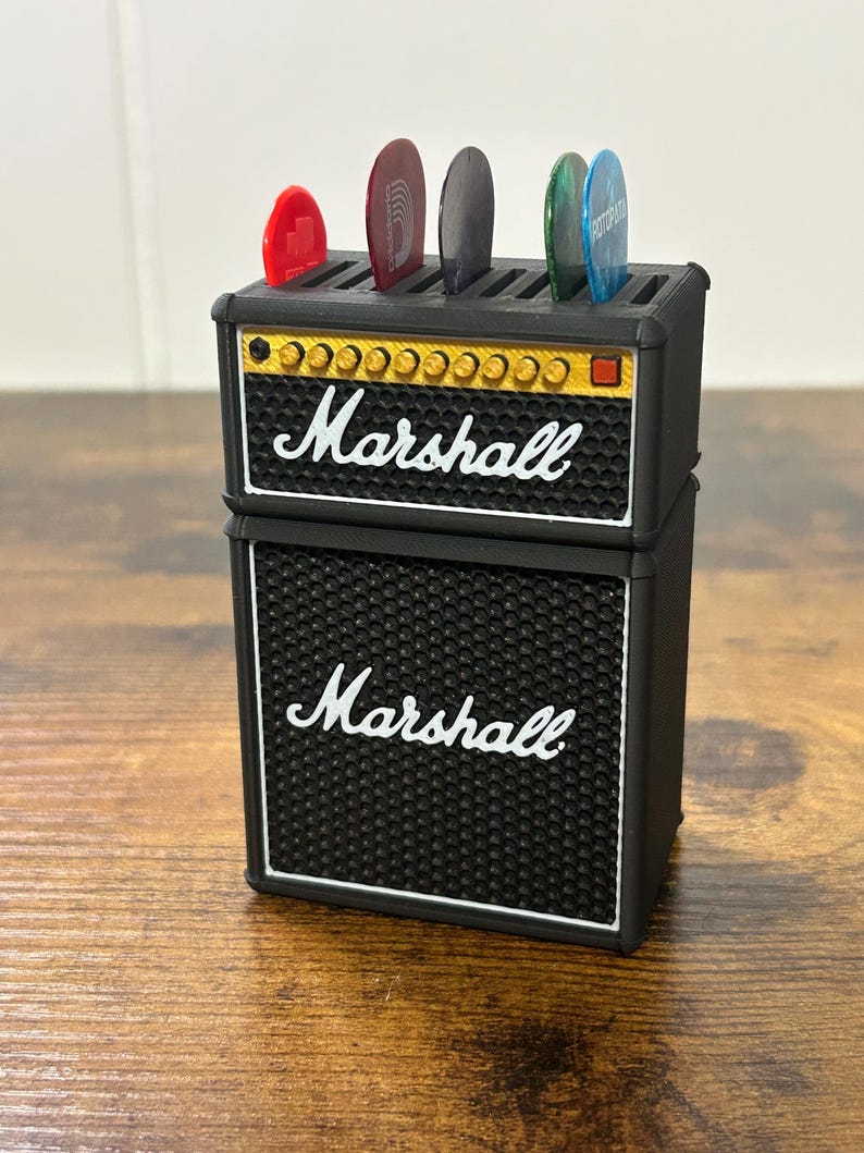 May include: Black and gold Marshall guitar pick holder shaped like a miniature amplifier. The holder has six slots for guitar picks and features the Marshall logo on the front.