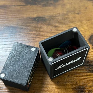 May include: Black guitar pick holder shaped like a Marshall amplifier. The lid is open and shows a variety of guitar picks inside.