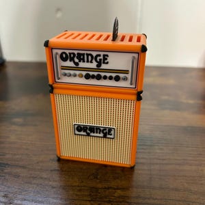 May include: Orange brand guitar amplifier with a black and orange colour scheme. The amplifier has a speaker grill and a control panel with knobs.