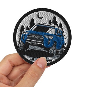 May include: A black and white embroidered patch featuring a blue Jeep driving through a forest at night. The patch includes a crescent moon and stars in the background.