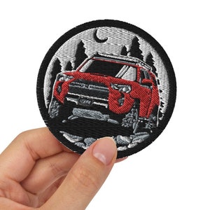 May include: A black and white embroidered patch depicting a red SUV driving through a forest at night. The patch features a crescent moon and stars in the background.