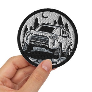 May include: A black and white embroidered patch depicting a white SUV driving through a forest at night. The patch features a moon and stars in the background.