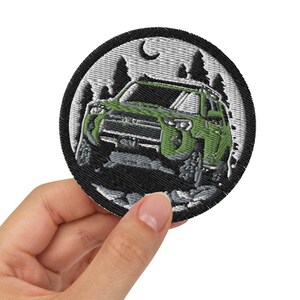 May include: A round embroidered patch with a black background and a white outline. The patch features a green SUV driving through a forest scene with a crescent moon in the sky.