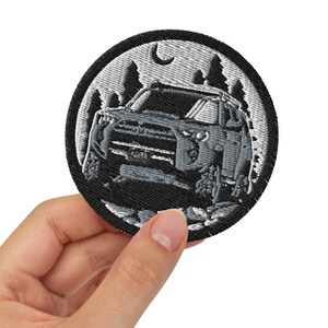 May include: A round, black and gray embroidered patch featuring a detailed off-road vehicle design. The patch includes a forest scene with trees and a crescent moon in the background. The patch is held in a hand.